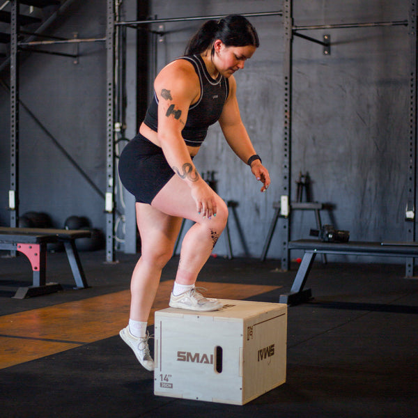 A person is performing a box step-up exercise on an SMAI Plyometric Box - Small Wood in a gym. They are dressed in a black tank top, black shorts, and white sneakers, with various gym equipment visible in the background.