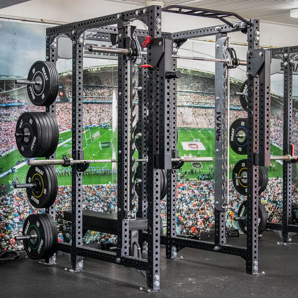 A power rack stands in the gym, brimming with weight plates on each side. Nearby, the SMAI Smith Machine Insert - Vanta Series awaits its turn. A mural of a stadium buzzing with spectators adds dynamism to the workout area, creating an invigorating ambiance for weightlifting enthusiasts.