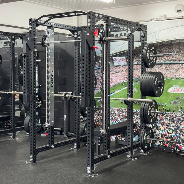 A gym scene showcases the SMAI Smith Machine Insert - Vanta Series, complete with barbells and weight plates, perfect for weightlifting. Positioned on a black floor, the machine is set against a large sports stadium mural in the background, capturing the energy of a football game and its cheering crowd.