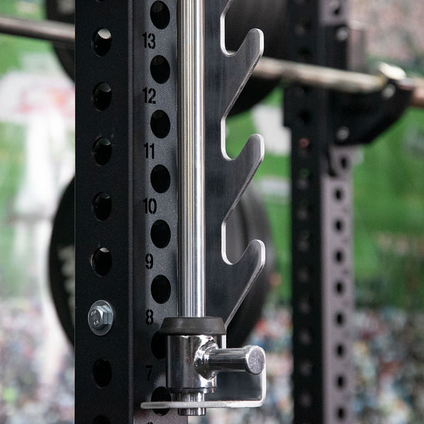 Close-up of the SMAI Smith Machine Insert from the Vanta Series in a gym, featuring numbered holes for securing pegs. The metal adjustable weight rack holds a barbell, set against a green and blurred background.