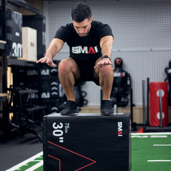 Wearing athletic attire, an individual is executing a box jump exercise in a gym, utilizing a durable SMAI Plyometric Box - Pre-Assembled marked "30 inches." The athlete is mid-air with arms extended forward for balance, ensuring safety. Various fitness equipment is visible in the background.