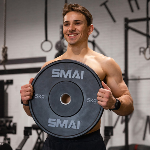 A man grins as he grips a pair of 5kg HD Bumper Plates from Sale Item in a gym, channeling the essence of Olympic lifting. He is shirtless, sporting a black wristwatch, and stands assuredly before gym equipment and rings.