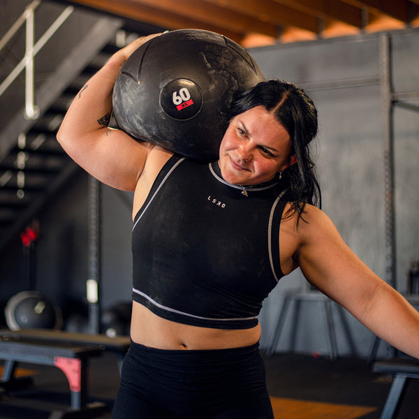 In a gym setting, Crossfit Athlete Julia Hannaford in a black sleeveless top lifts a 60 kg SMAI Slam Ball Monster from the set—which includes 60 kg, 70 kg, and 80 kg options—onto their shoulder. This durable rubber equipment is ideal for strongman training, with various workout tools visible in the background.