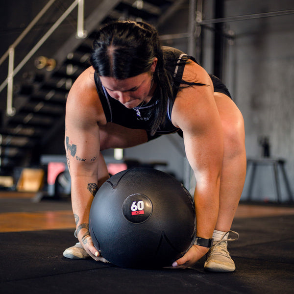 Crossfit Athlete Julia Hannaford lifts a 60 kg Slam Ball from the SMAI Slam Ball Monster Set, perfect for strongman training, in a gym. They are focused on the exercise, with tattoos visible on their arms. The space features a dark interior with durable rubber gym equipment and a staircase in the background.