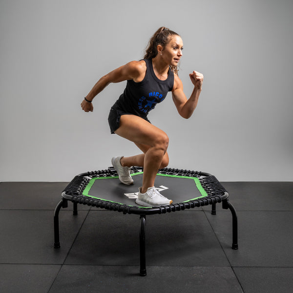 Jogging on the 50" Rebounder Mini Trampoline by SMAI, a woman in a black tank top and shorts enjoys her workout with sturdy bungee cords against a plain gray backdrop.