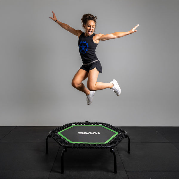 A woman in athletic attire jumps on a 50" SMAI Rebounder Mini Trampoline, arms outstretched. She's in a black tank top with shorts and white sneakers. The trampoline's hexagonal shape adds flair to the plain gray background.