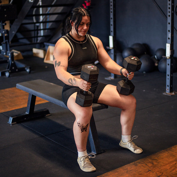 A person with tattoos sits on a bench in a gym, clutching an SMAI Rubber Hex Dumbbell - (Pair) Apex in each hand. They're dressed in a black tank top and shorts, paired with white sneakers. Premium quality materials are evident throughout the gym equipment visible in the background.