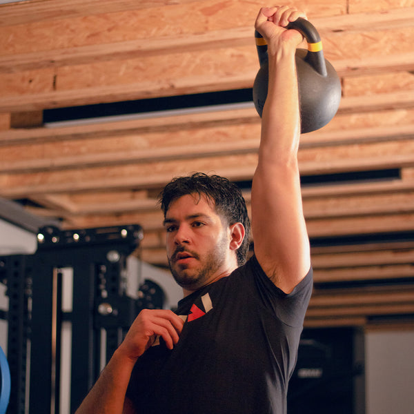 Amidst a gym filled with equipment, an individual lifts a SMAI Competition Steel Kettlebell - Black overhead using one arm. They stand focused, clad in a black shirt, under the wooden ceiling beams of the room.