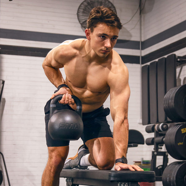 A person working out in a gym performs a one-arm row using the SMAI Competition Steel Kettlebell - Black while seated on a bench. Shirtless, wearing shorts and a watch, they maintain a focused expression. The kettlebell, distinguished by its color identifier rings, is part of the visible gym equipment in the background.