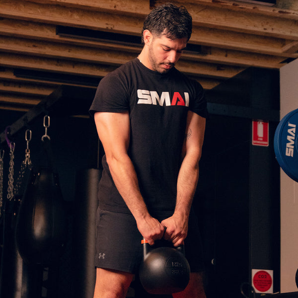 In a gym, a man in black attire exercises with a SMAI Competition Steel Kettlebell - Black. He concentrates on the weight, its color identifier rings subtly standing out against the dimly lit surroundings, while punching bags loom in the background.