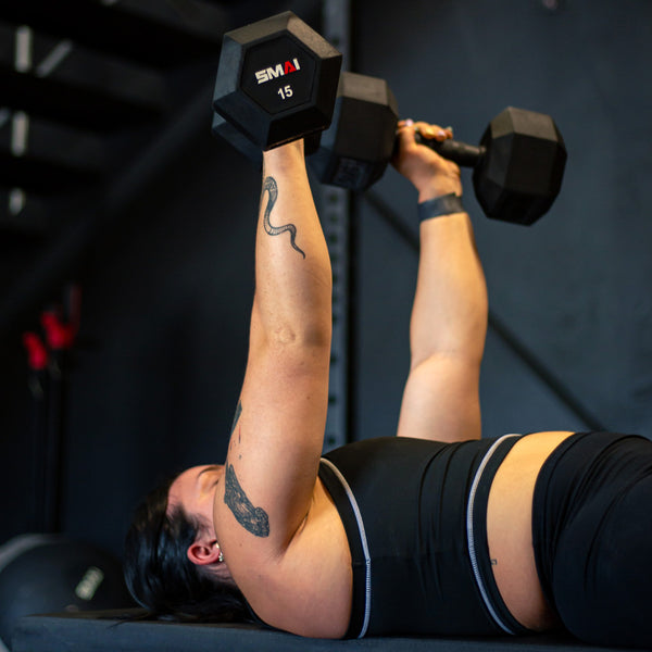 Clad in athletic gear, a person is reclining on a bench and hoisting the SMAI Rubber Hex Dumbbells - (Pair) Apex above their chest. A snake tattoo adorns their arm. The environment resembles a gym with dark walls and various exercise equipment, creating an ideal atmosphere for an intense fitness session.