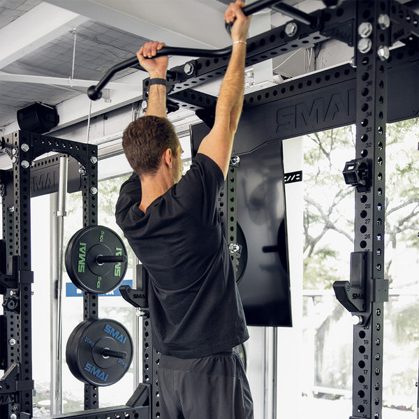 A man in a black shirt and gray shorts performs pull-ups on the SMAI Double Sided Power Rack - Vanta Series. The bright room reveals trees through the windows, while weight plates are neatly stacked on the rack beside him.