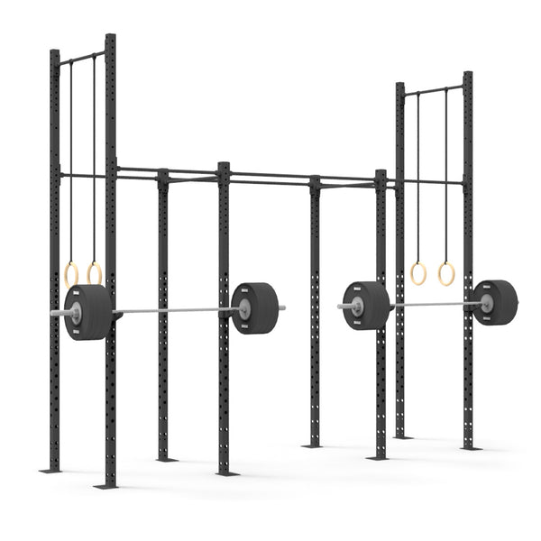 The gym setup features the Rig - 4 Squat Cell with Bridge by No Sync To Channel Engine, constructed from durable steel tubing and equipped with loaded barbells and hanging gymnastic rings. This commercial equipment is displayed against a plain white background, accentuating its robust modular design.