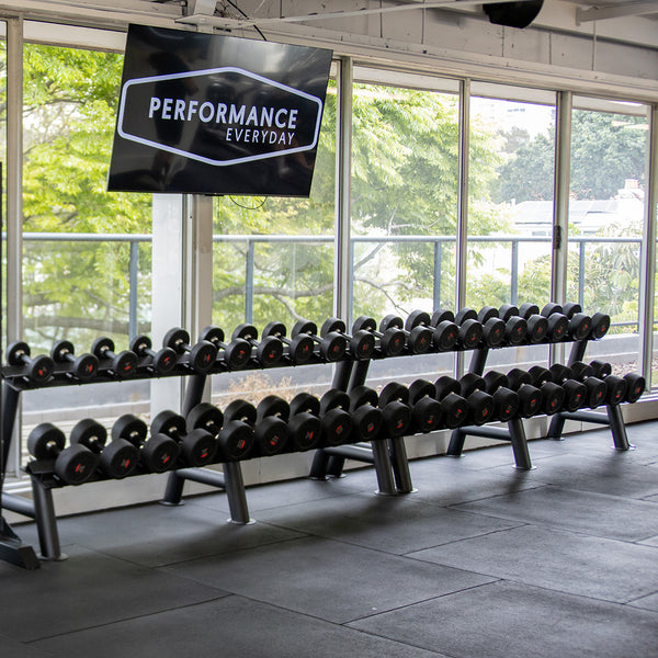 A gym interior showcasing the SMAI Commercial Dumbbell Set with Rack 32.5-60kg, neatly arranged on a sleek storage rack positioned in front of large windows that offer a picturesque view of the trees outside. Above, a sign proclaims "Performance Every Day." The gym exudes a clean and organized look, complete with a rubber mat floor.