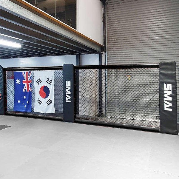 The SMAI MMA Cage Panel Pack includes a robust chain-link fence, sturdy padding with reinforced mesh for safety, and is adorned with flags from Australia, the UK (partially visible), and South Korea. Grey mats are used to cover the floor.